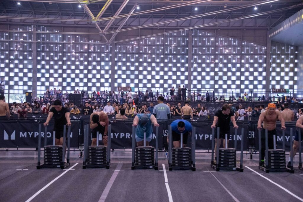 Was ist Hyrox: Athleten schieben beim Wettkampf beschwerte Schlitten in der Halle